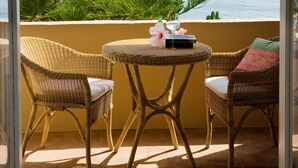 Un balcon avec des fauteuils en osier, une petite table avec un livre et un verre, donnant sur la mer, avec des bateaux et une végétation luxuriante.