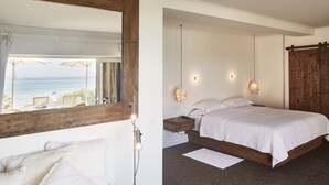 Une chambre à coucher avec un grand lit, une tête de lit en bois, une table de chevet et une commode. Il y a une fenêtre donnant sur l’océan et une plage.