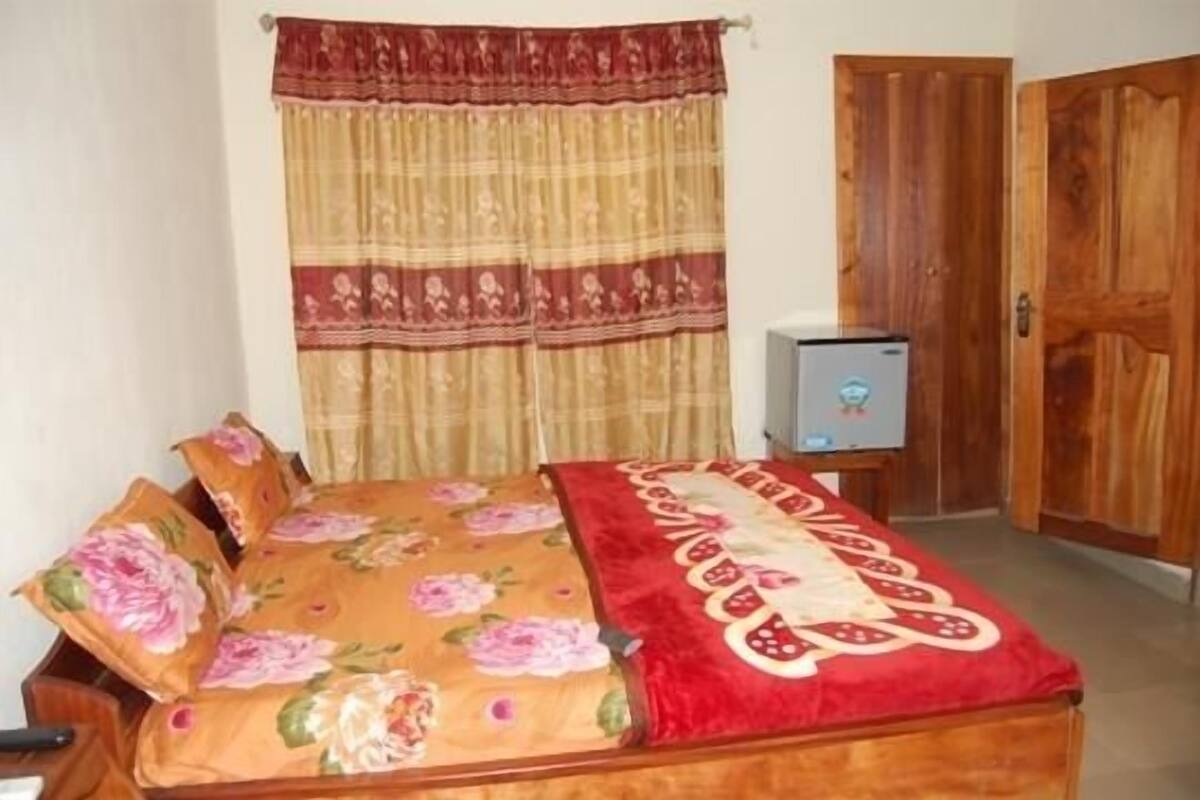 A bedroom with a bed, curtains, a wooden wardrobe, and a small table with a television.