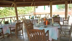 An outdoor dining area with wooden tables and chairs, covered by a wooden roof, and surrounded by greenery.