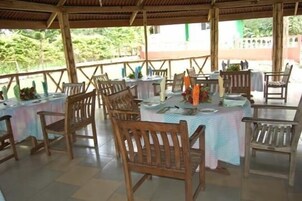 An outdoor dining area with wooden tables and chairs, covered by a wooden roof, and surrounded by greenery.