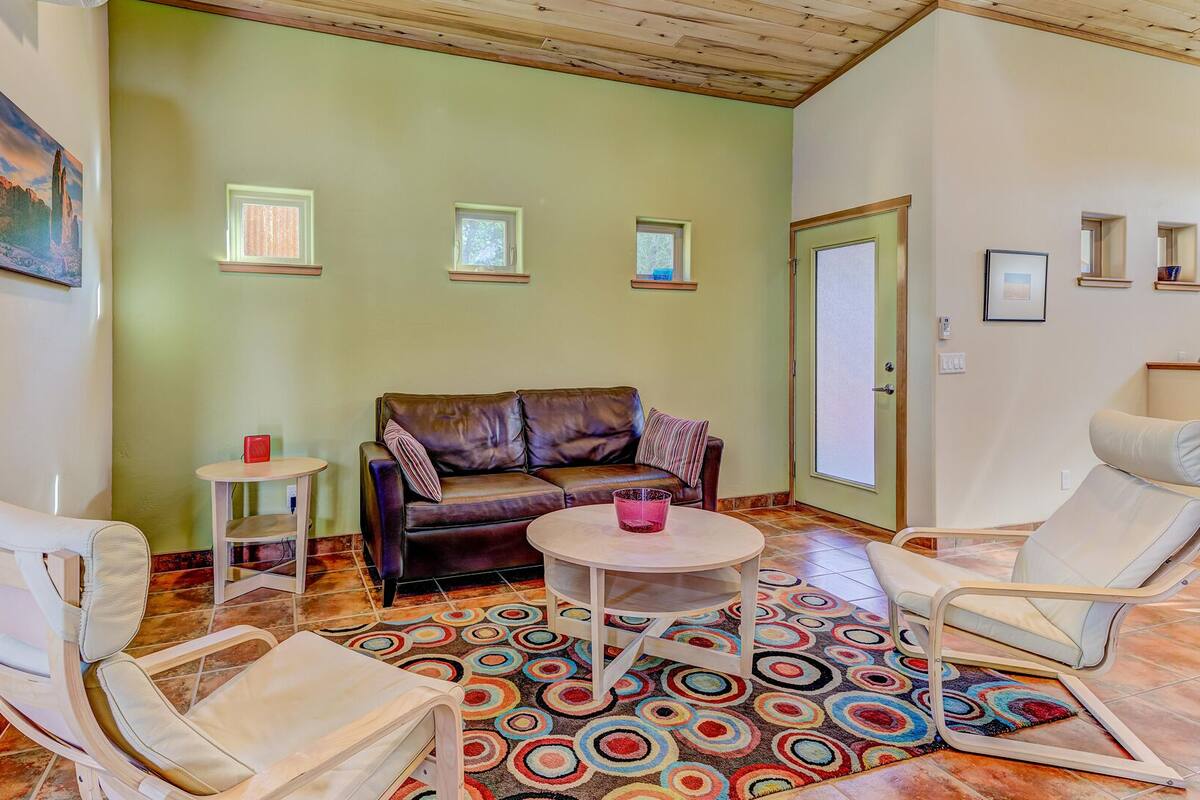 A living room with a brown leather sofa, two white armchairs, a small round table, and a colorful circular rug. There are three small windows and a door with frosted glass.
