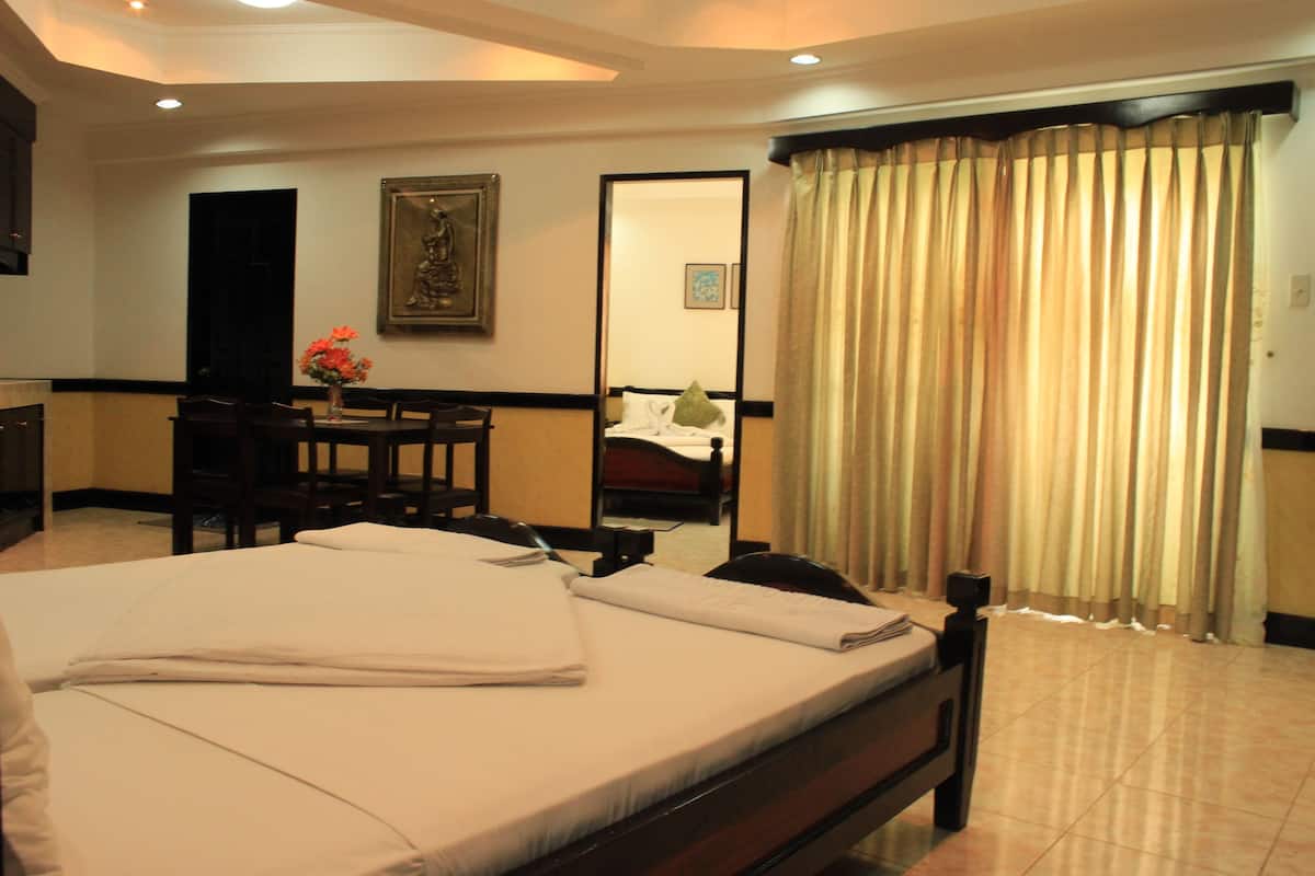 A hotel room with a neatly made bed, a desk with a chair, a dining table with chairs, and a large window with curtains.