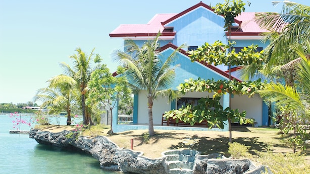 Une propriété située en bord de mer, avec un bâtiment bleu, des palmiers et un chemin de pierre menant à l’eau.