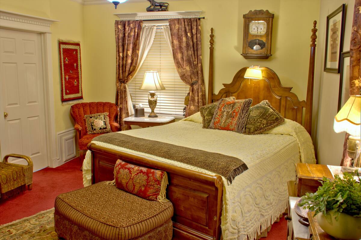 A traditional bedroom with a wooden bed, a red armchair, a small table with a lamp, and a clock on the wall.