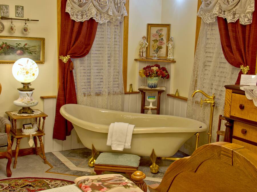 A vintage bathroom with a clawfoot bathtub, a wooden vanity, a small table with a lamp, and a floral rug.