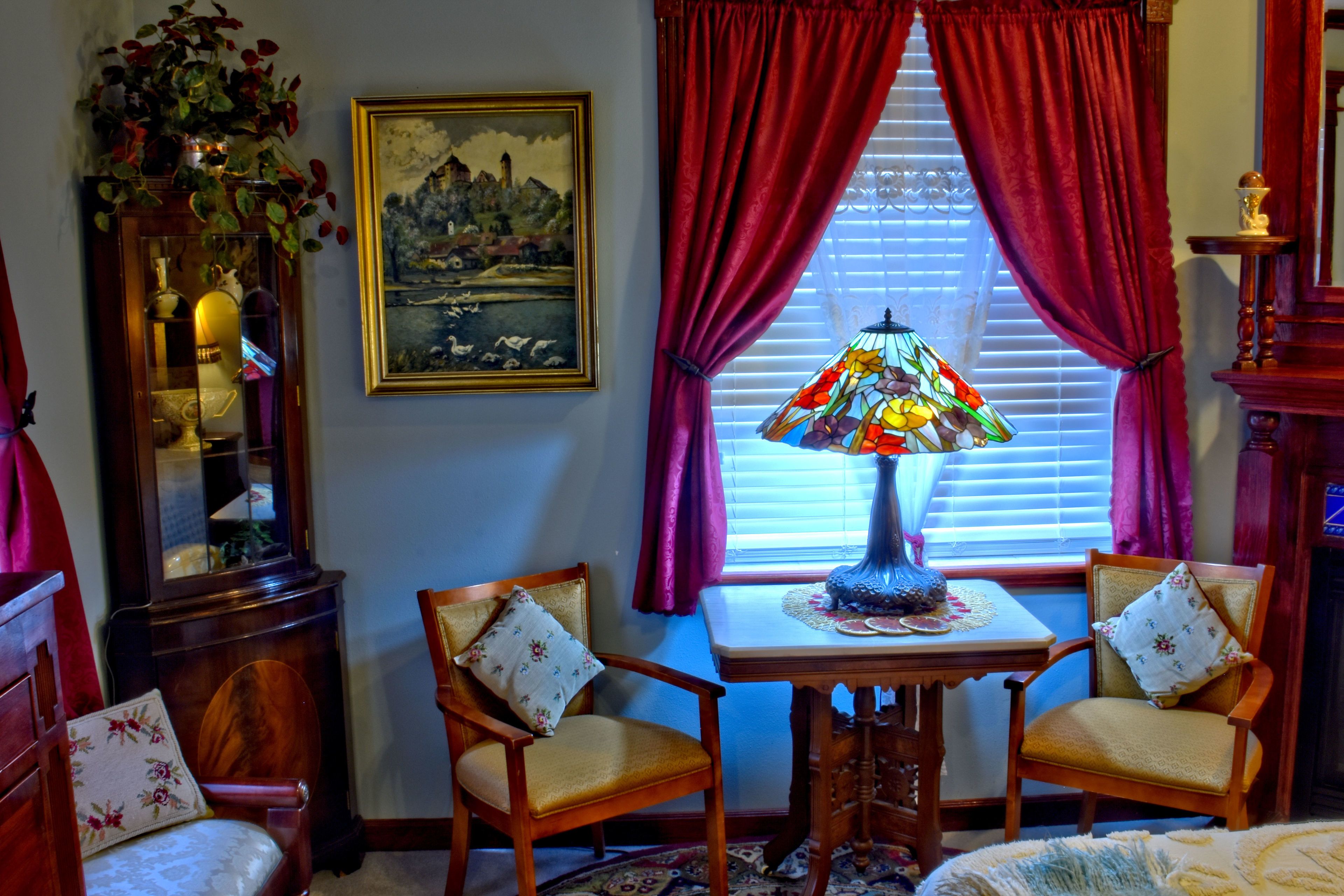A room with a wooden table, two chairs, a stained glass lamp, a painting, and a window with red curtains.