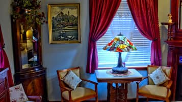 A room with a wooden table, two chairs, a stained glass lamp, a painting, and a window with red curtains.