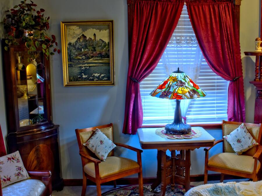 A room with a wooden table, two chairs, a stained glass lamp, a painting, and a window with red curtains.