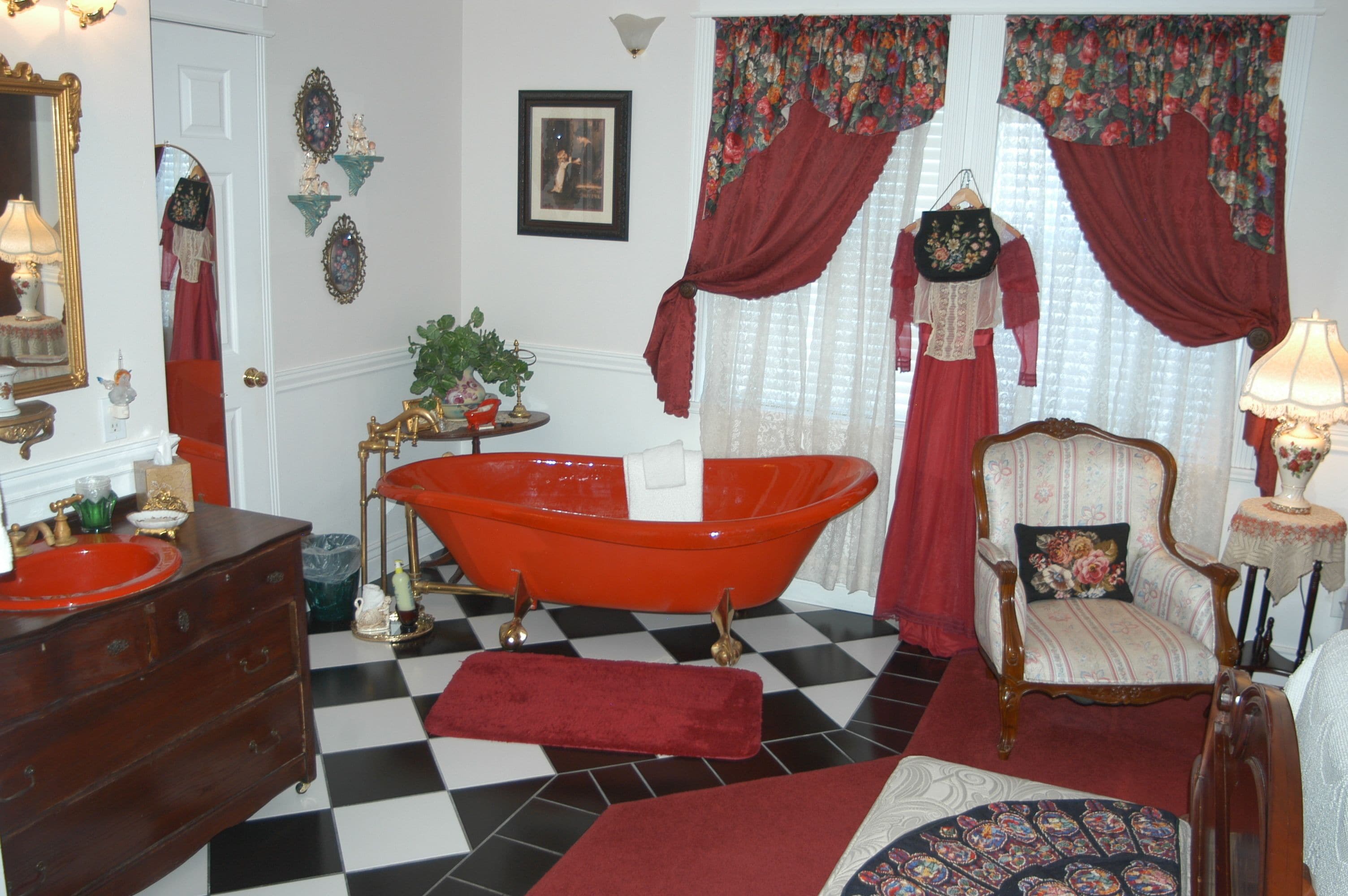 A vintage bathroom with a checkered floor, a red bathtub, a wooden dresser, and a chair with a floral cushion.