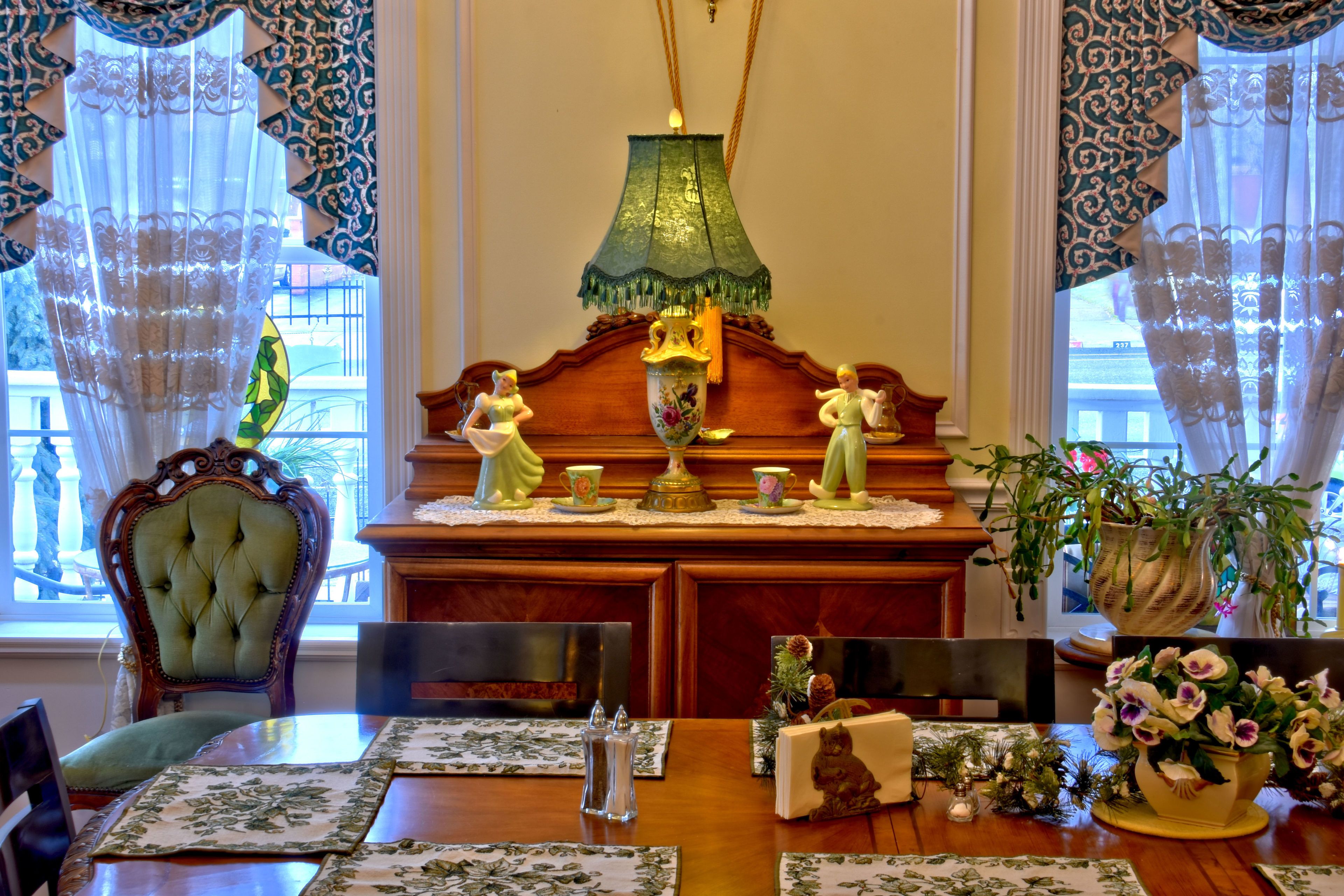 A dining area with a wooden table set with floral placemats, a green table lamp, and a vase with flowers.