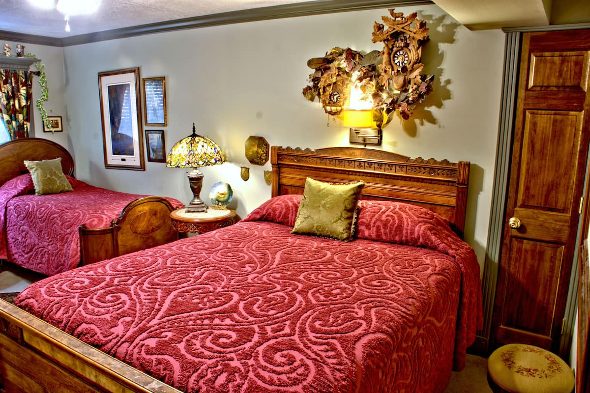 A room with two wooden beds, each with a red patterned bedspread and matching pillows. There is a nightstand with a lamp and a wall-mounted clock.