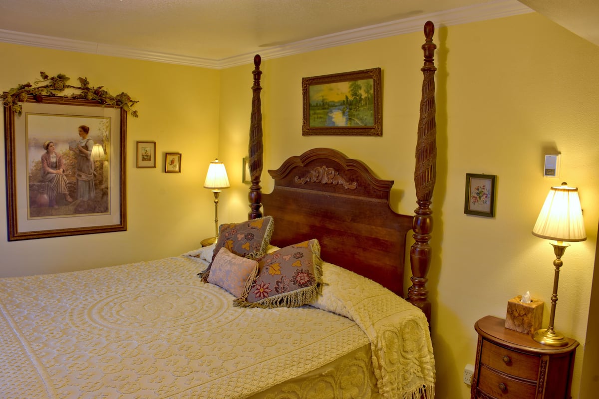 A four-poster bed with floral pillows, a nightstand with a lamp, and a painting on the wall.