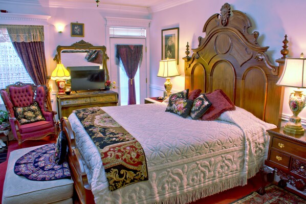 A classic bedroom with a large bed, a wooden headboard, a red armchair, and a mirror above the dresser.