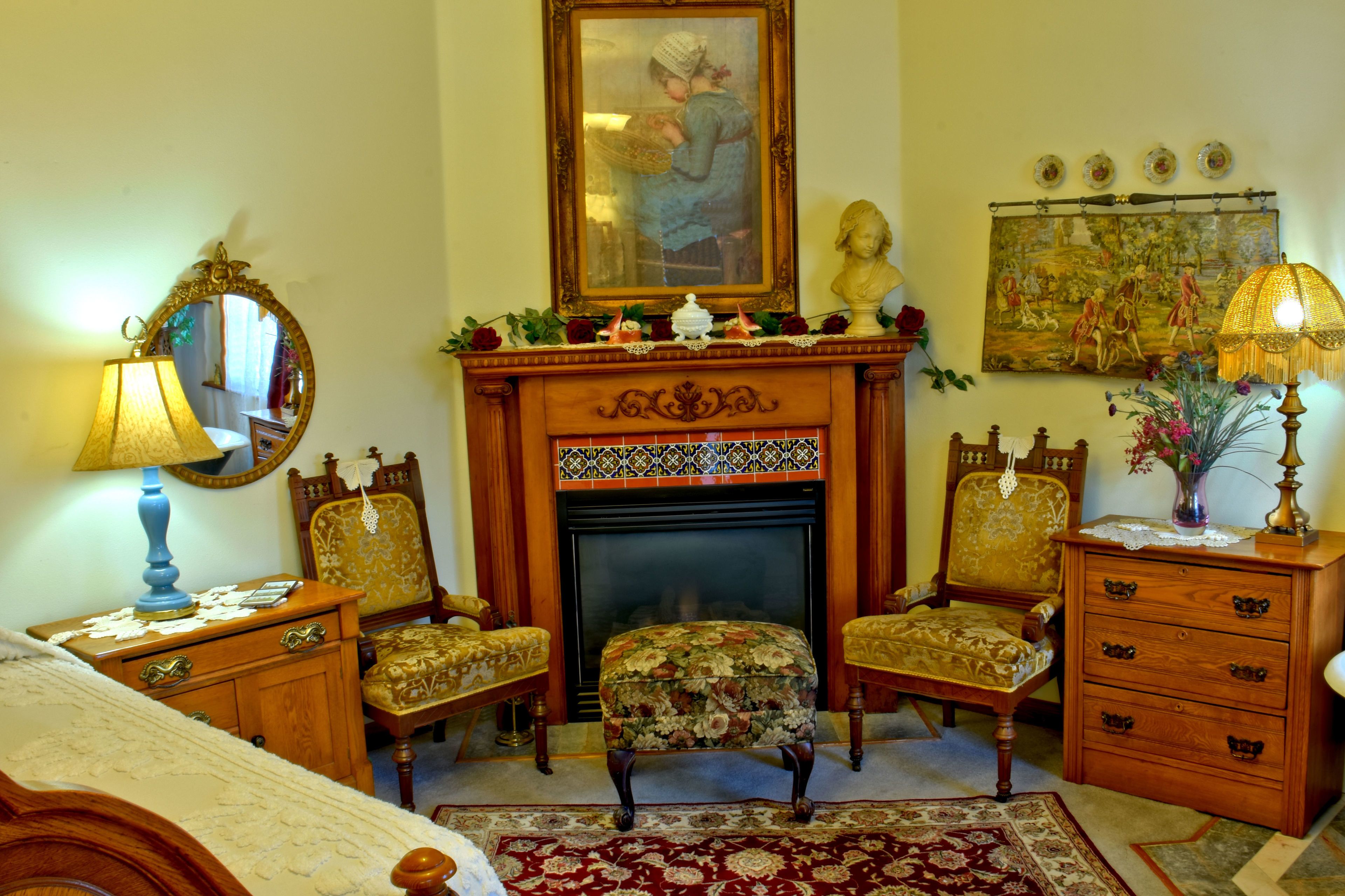 A traditional room with a fireplace, two upholstered chairs, a wooden dresser, and a bed.
