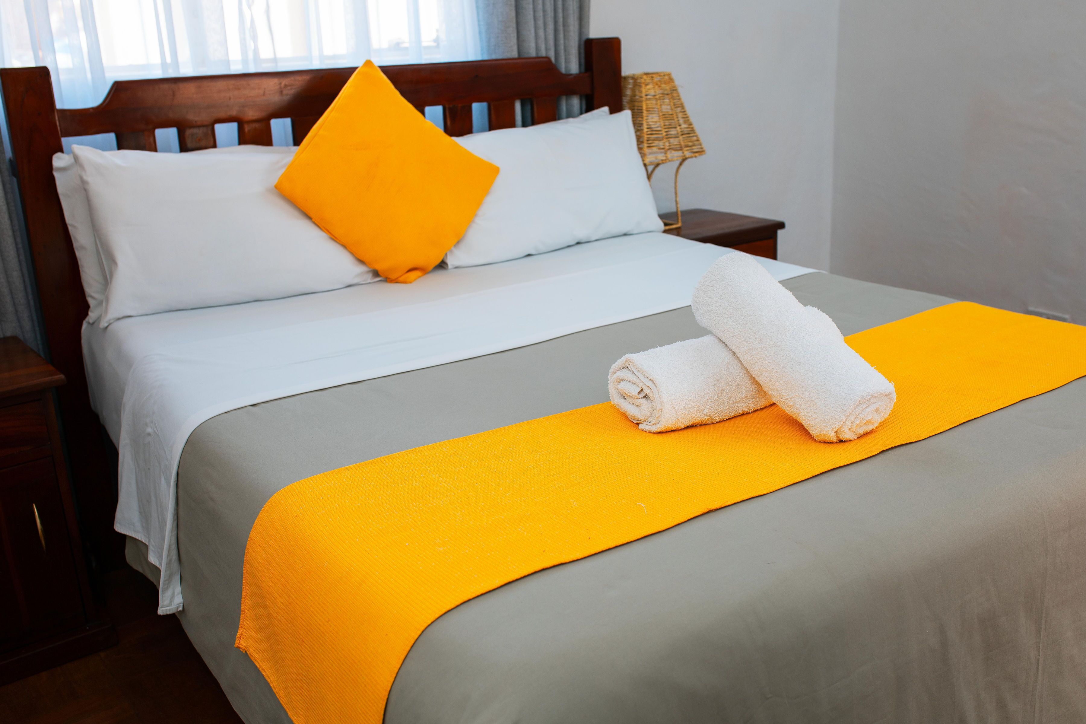 A neatly made bed with white and gray bedding, a bright orange pillow, and two rolled towels on a matching orange blanket.