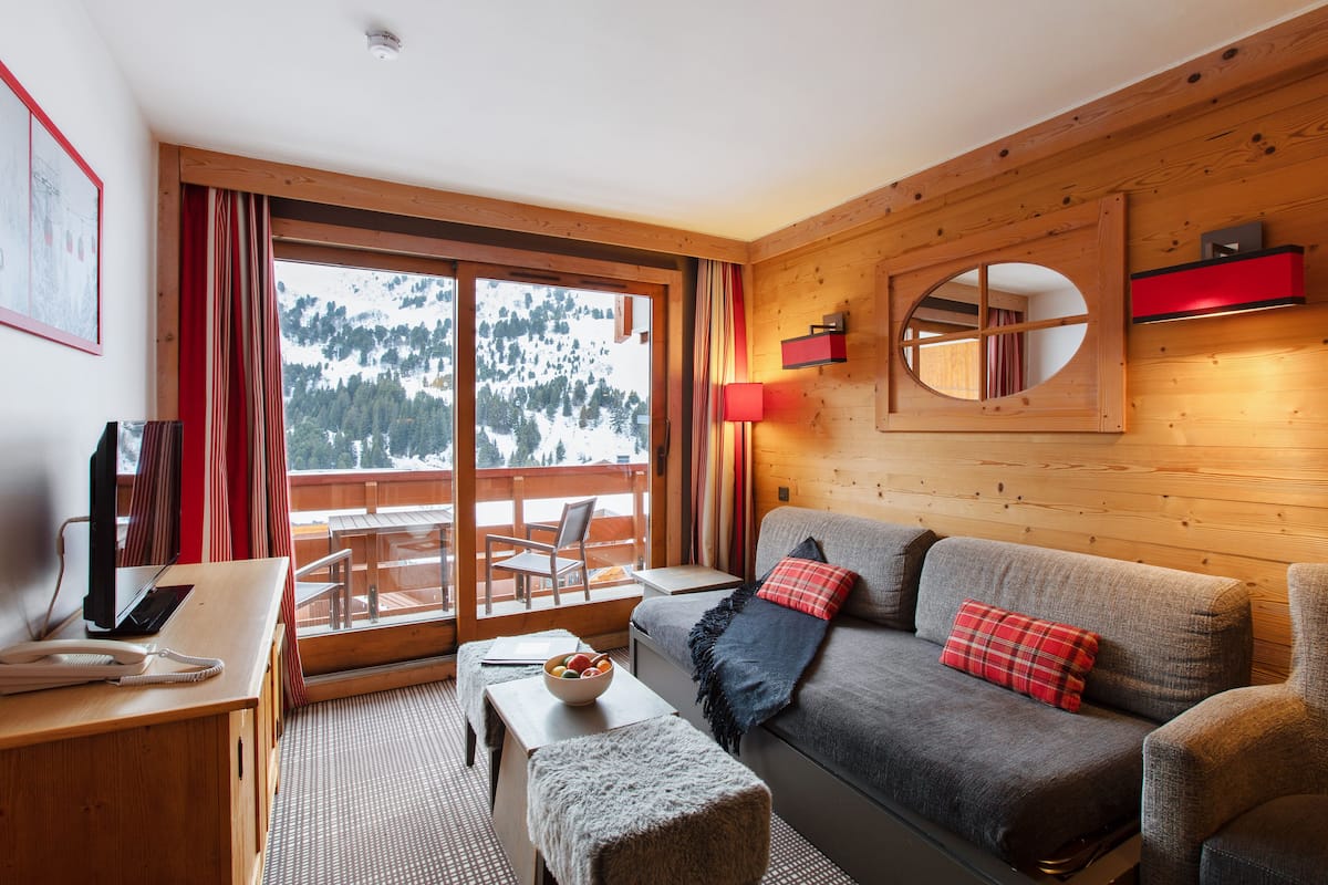 Une pièce chaleureuse avec un décor intérieur en bois, un canapé, une petite table et une vue sur des montagnes enneigées par de grandes fenêtres.