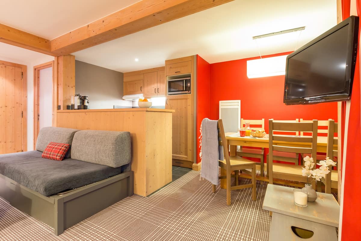 Une pièce compacte comprenant un canapé, une table à manger avec des chaises, un téléviseur fixé au mur et un coin cuisine.