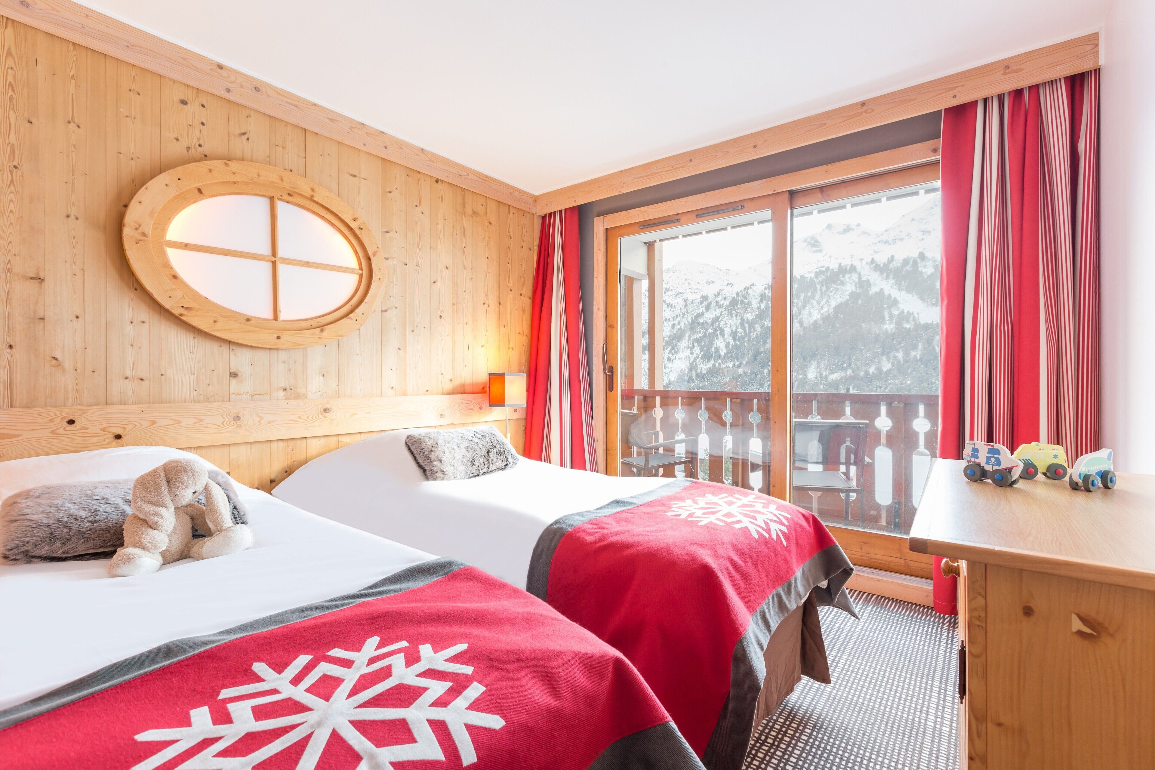 A room with two beds, a wooden headboard, a window with curtains, a view of mountains, and a small table with toys.