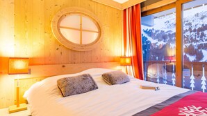 Une chambre d’hôtel avec un intérieur en bois, un grand lit avec une literie blanche, deux oreillers, un plaid rouge et une fenêtre donnant sur des arbres couverts de neige.