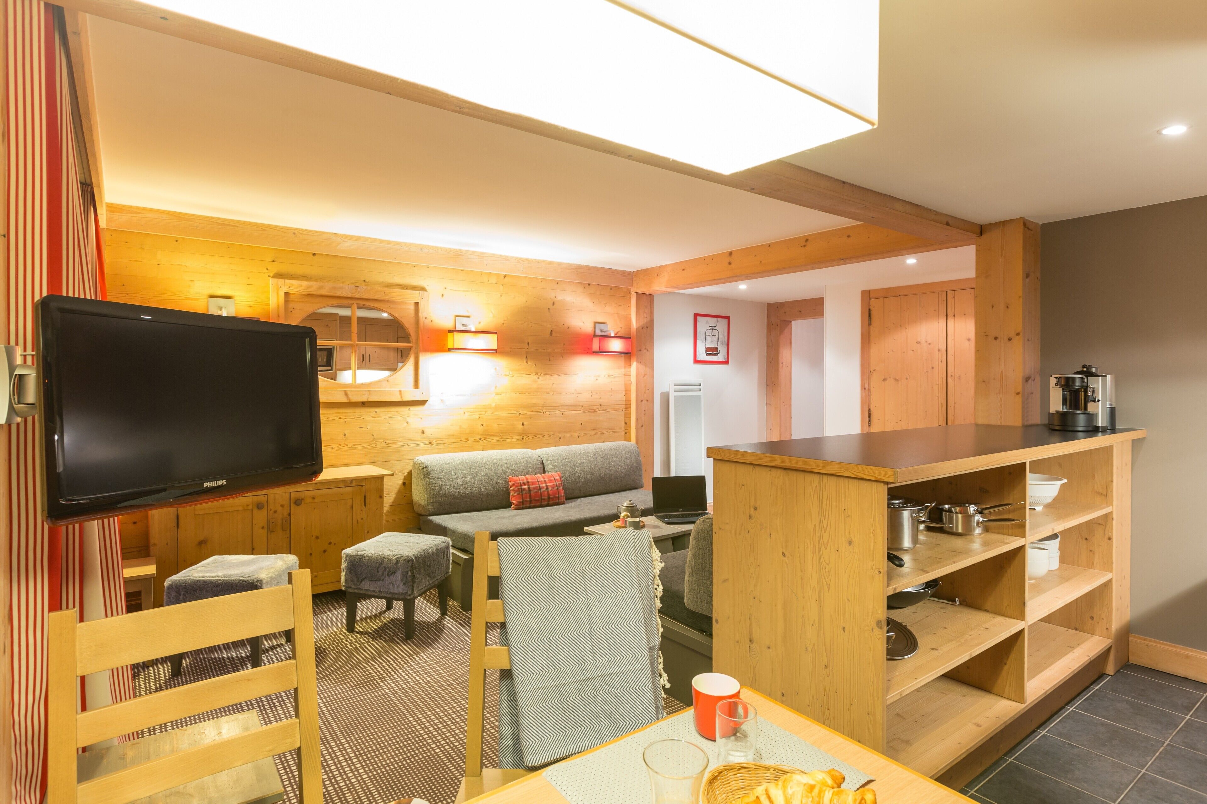 A cozy room with a wooden ceiling, a flat-screen TV, a sofa, a small table with a cup and a basket, and a kitchen area with wooden cabinets and a coffee maker.