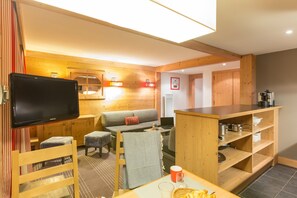 Ein gemütliches Zimmer mit Holzbalkendecke, einem Flachbildfernseher, einer Couch, einem kleinen Tisch mit Tasse und Korb sowie einer Küchenzeile mit Holzausstattung und einer Kaffeemaschine.