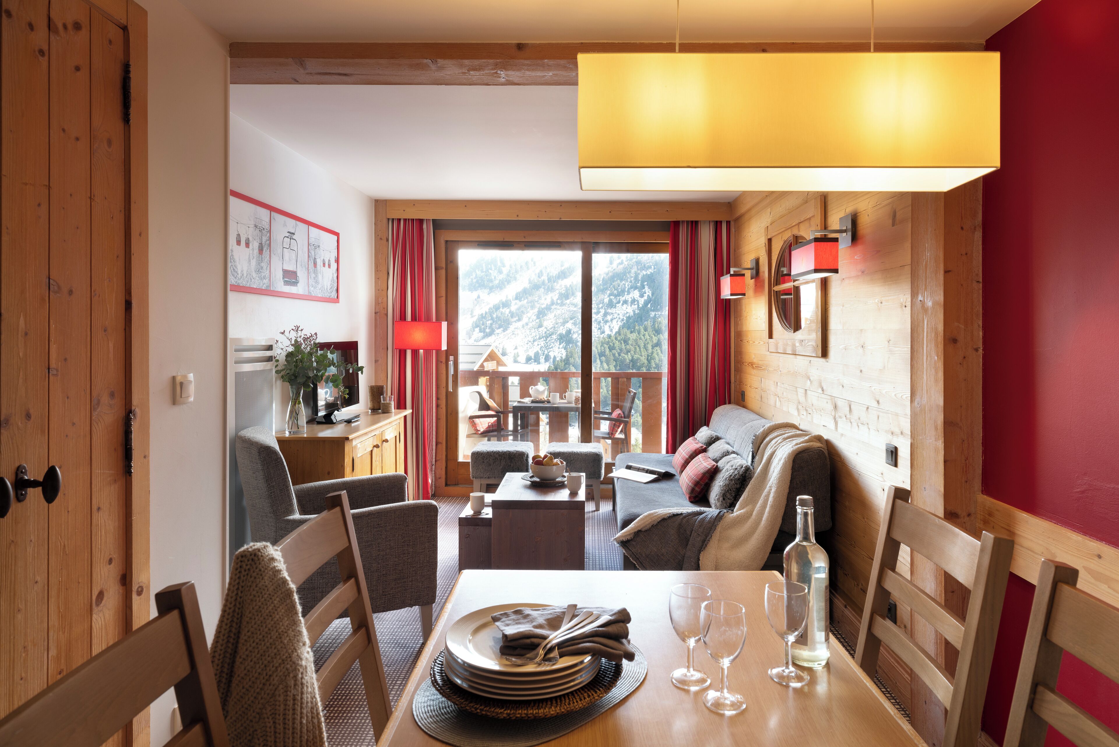 Un coin repas chaleureux avec une table en bois dressée pour deux personnes, un canapé et une vue sur une montagne enneigée.