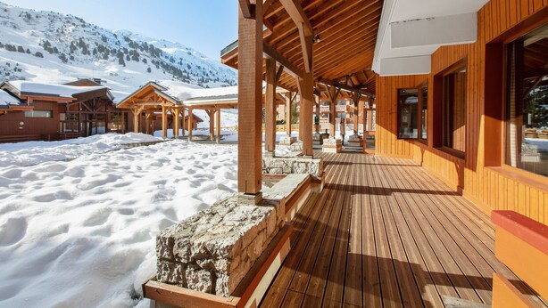 Une terrasse en bois, le sol recouvert de neige, des bâtiments en bois dotés de grandes fenêtres et un paysage montagneux en arrière-plan.