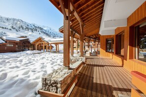 Une terrasse en bois, le sol recouvert de neige, des bâtiments en bois dotés de grandes fenêtres et un paysage montagneux en arrière-plan.