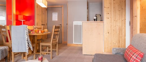 A cozy living area with a sofa, a table with a book and glasses, a wooden dining table with chairs, and a kitchen area with a refrigerator and a coffee machine.