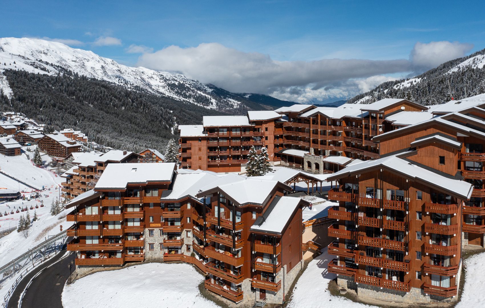 A snow-covered mountain resort with wooden buildings featuring balconies and ski slopes in the background.