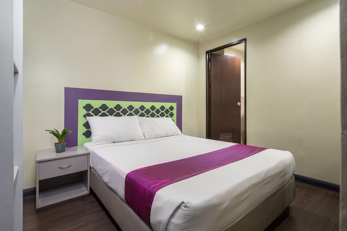 Un lit bien fait, recouvert d’une couvre-lit violette, une table de chevet avec une plante et une tête de lit ornée d’un motif géométrique.