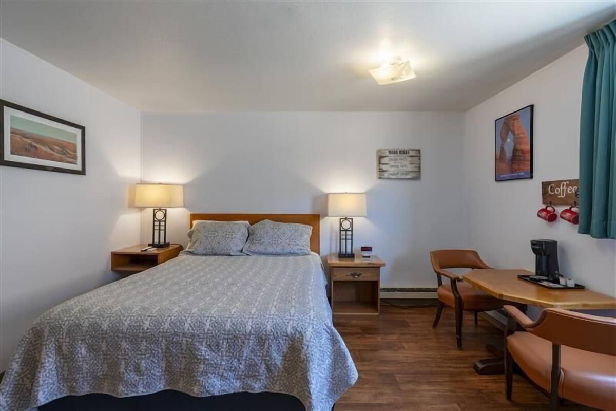 A bedroom with a bed, two bedside lamps, a wooden nightstand, a desk with a chair, and a coffee machine.