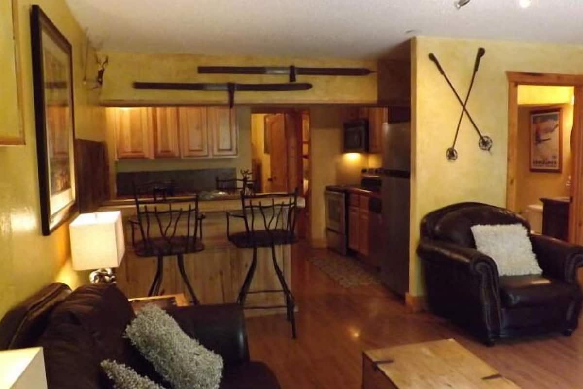 Una sala de estar con un sofá de cuero marrón, un sillón de cuero negro y una zona de comedor con un taburete de bar y armarios de madera.