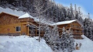 Una cabaña de madera con techo nevado, rodeada de árboles y un paisaje cubierto de nieve.