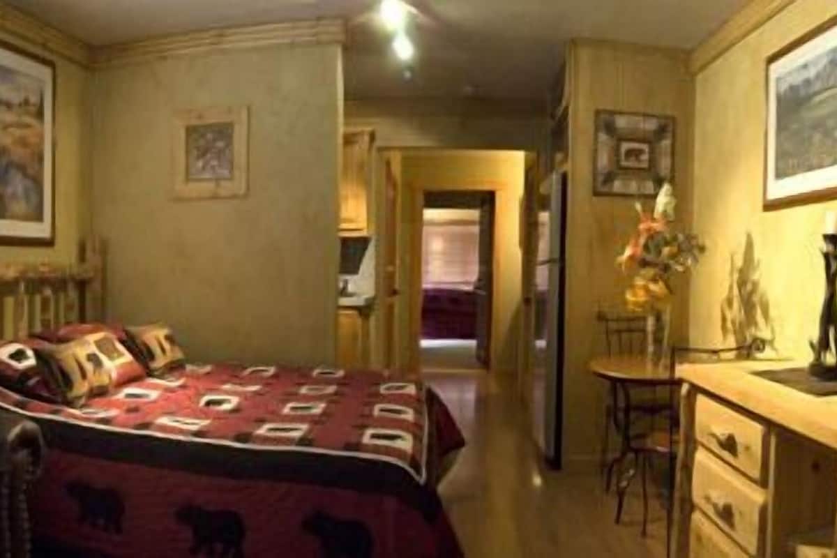 Una habitación con cama, sofá, escritorio y silla. Hay cuadros en las paredes y una ventana con persianas.
