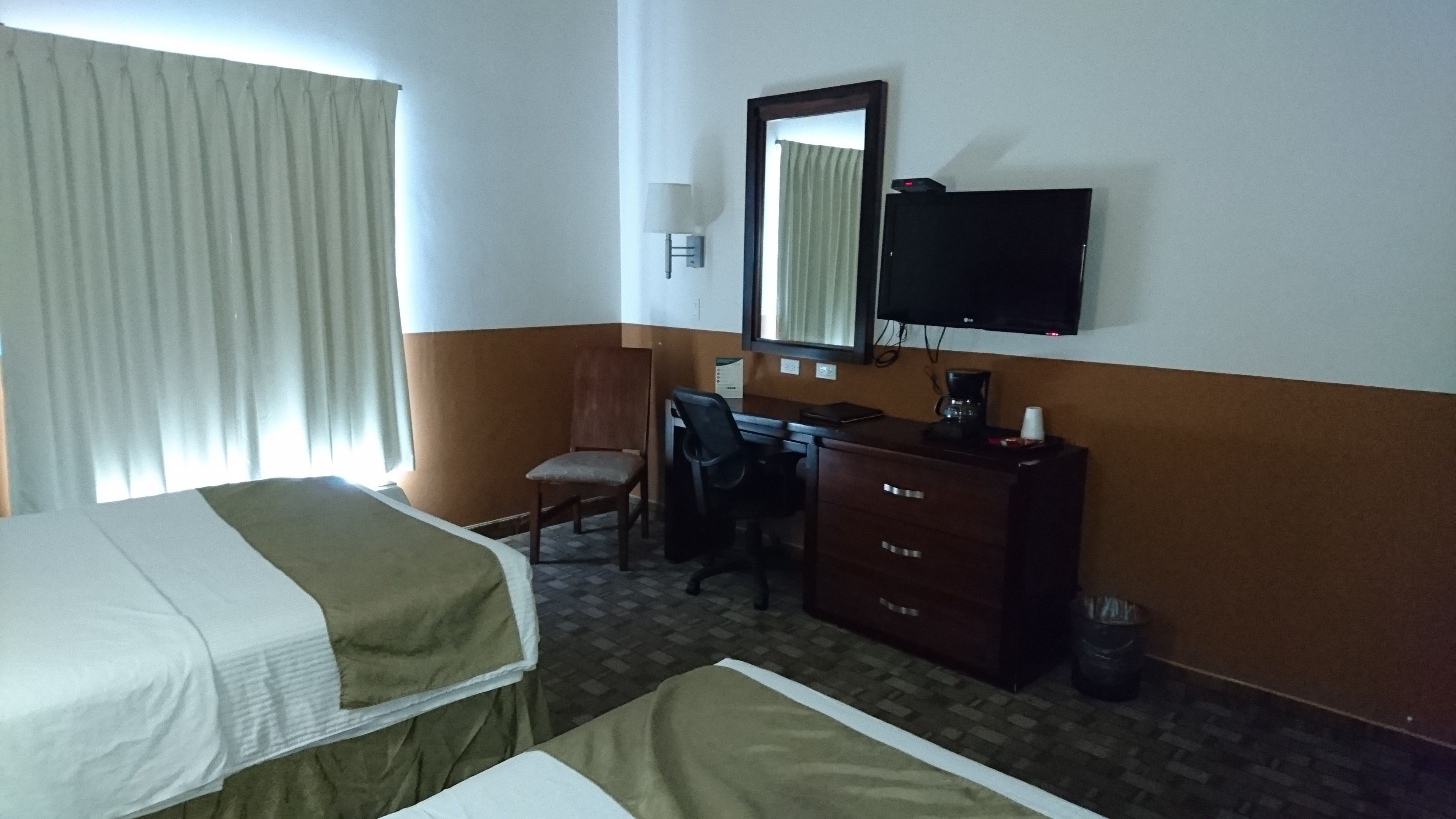 Ein Hotelzimmer mit einem Bett, einem Schreibtisch, einem Stuhl, einer Kommode, einem Fernseher und einem Fenster mit Vorhängen.
