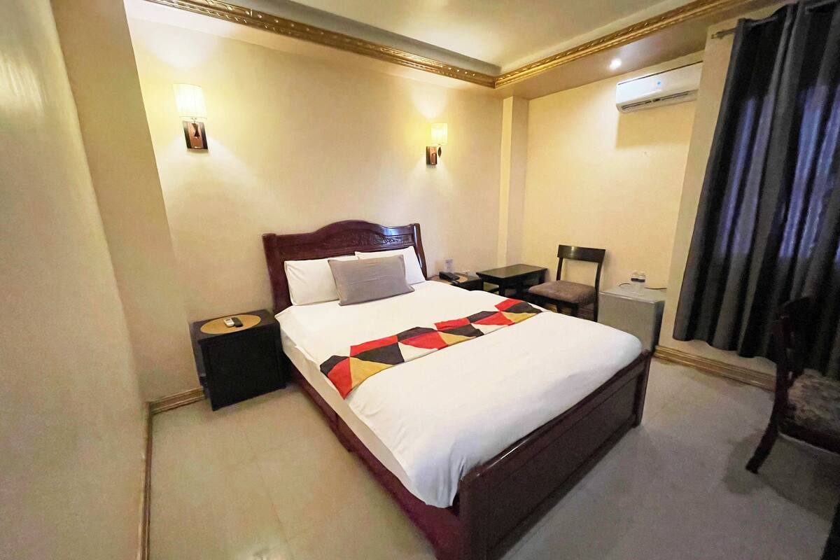 A hotel room with a large bed, bedside tables, a desk, and an air conditioning unit.