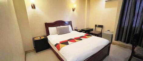 A hotel room with a large bed, bedside tables, a desk, and an air conditioning unit.