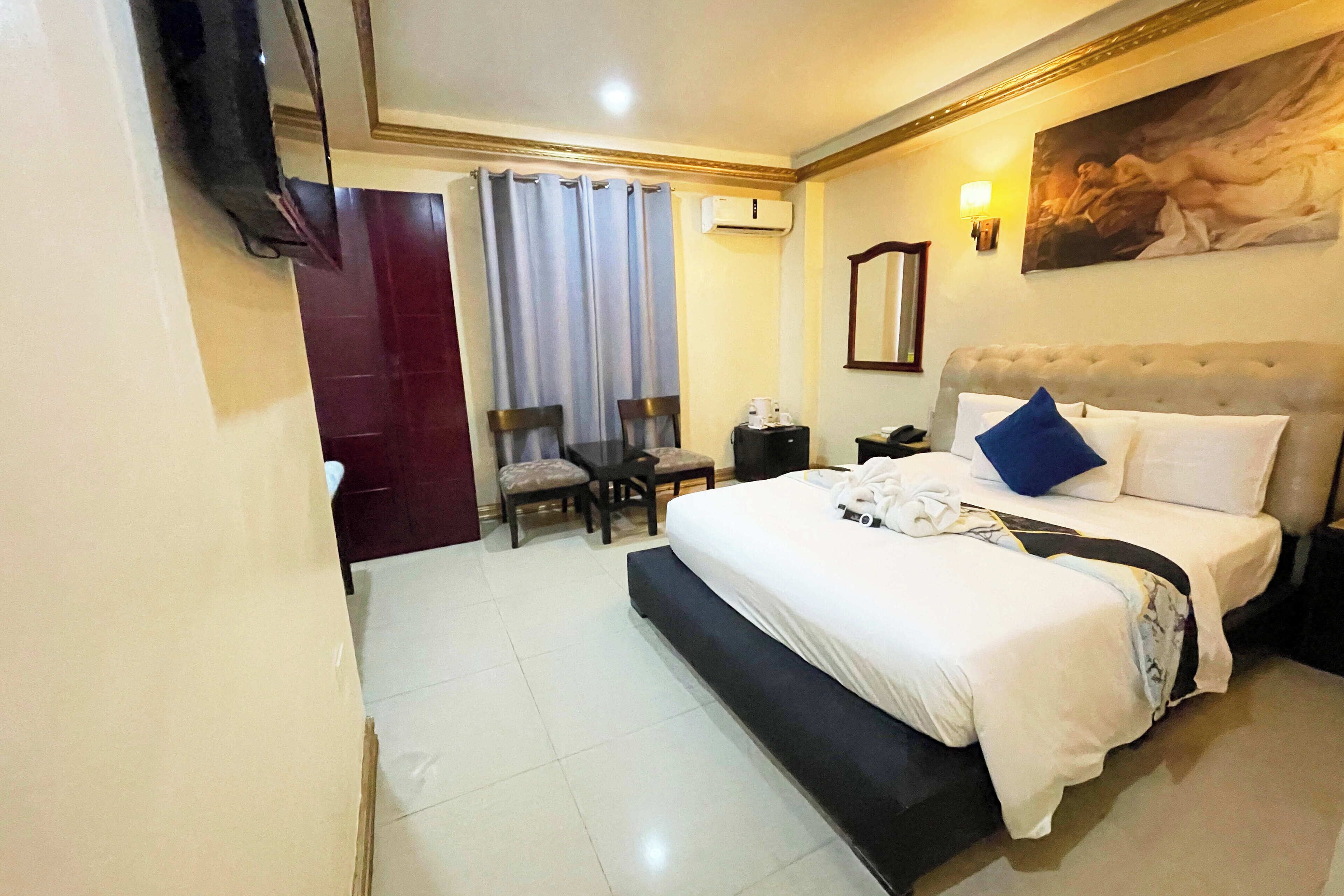 A hotel room with a large bed, a desk with two chairs, a flat-screen TV mounted on the wall, and a painting above the bed.