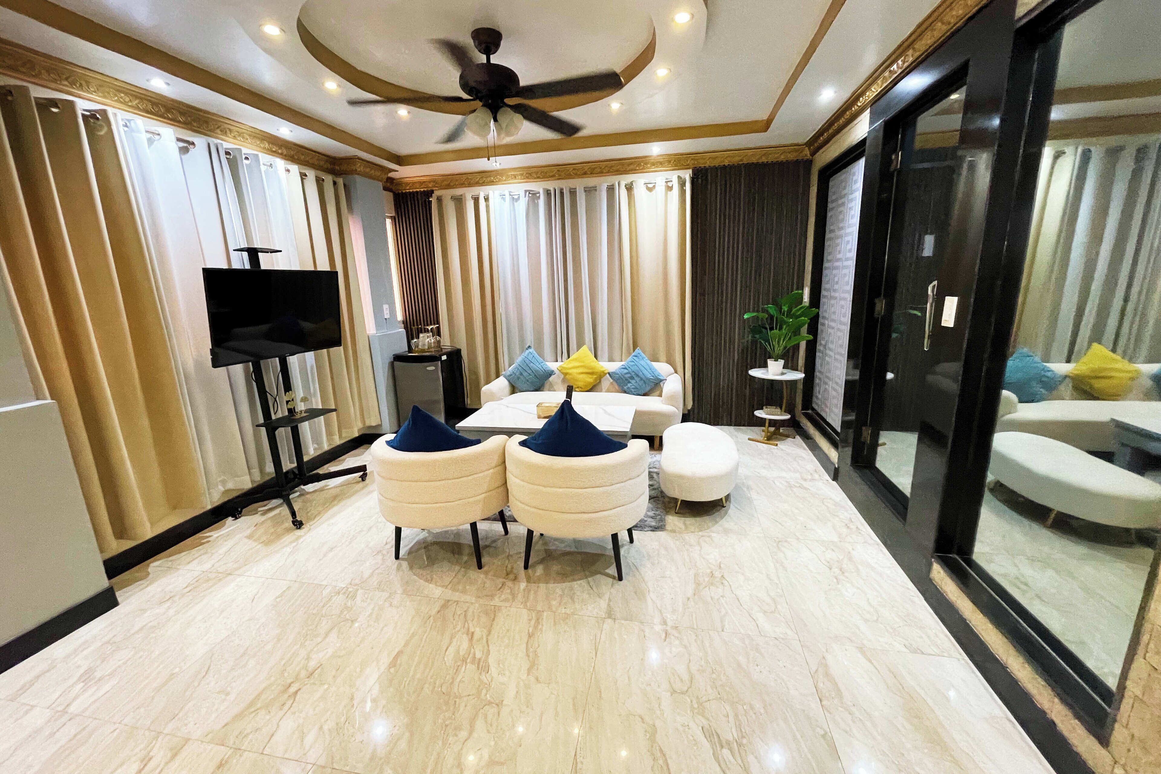 A modern living room with a flat-screen TV, a ceiling fan, a coffee table, and seating with cushions.