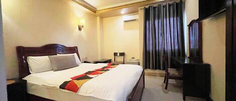 A hotel room with a large bed, bedside tables, a desk, a chair, a TV, and a curtain.