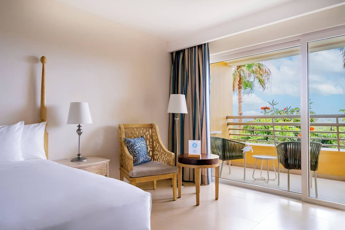 A hotel room with a bed, a chair, a table, and a balcony with a view of the ocean and palm trees.