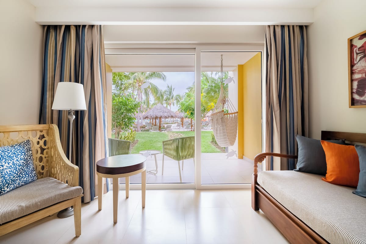 A hotel room with a wooden bed, a sofa, a small table, and a view of a garden with a thatched-roof structure.