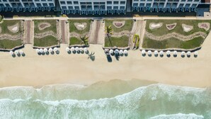 An aerial view of a beachside hotel with rows of beach chairs and umbrellas, a palm tree, and a wave crashing onto the shore. - Iberostar Selection Playa Mita - All Inclusive (Punta de Mita)