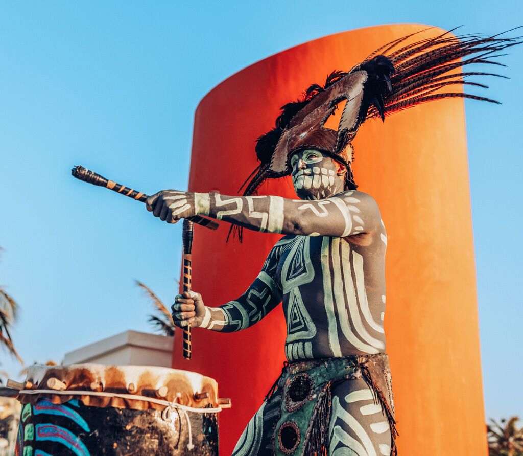 Une statue représentant une personne tenant un bâton, vêtue d’une tenue à motifs et coiffée d’un couvre-chef orné de plumes, le tout sur un fond rouge.