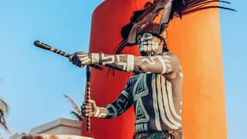 A statue of a person holding a staff, wearing a patterned outfit and a feathered headdress, set against a red background.