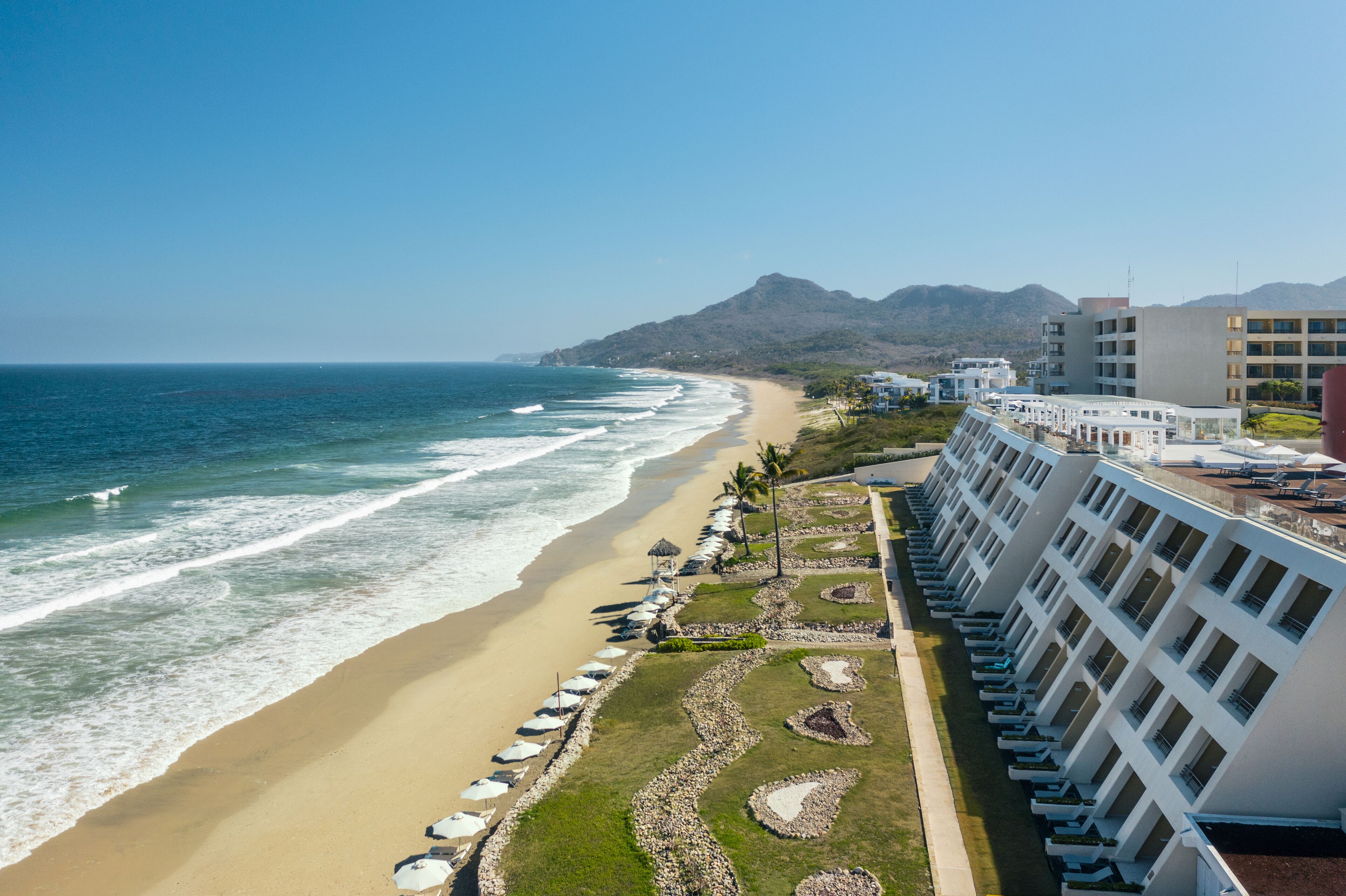 A beachfront hotel with a long row of rooms overlooking the ocean, a sandy beach, and a mountain in the background.