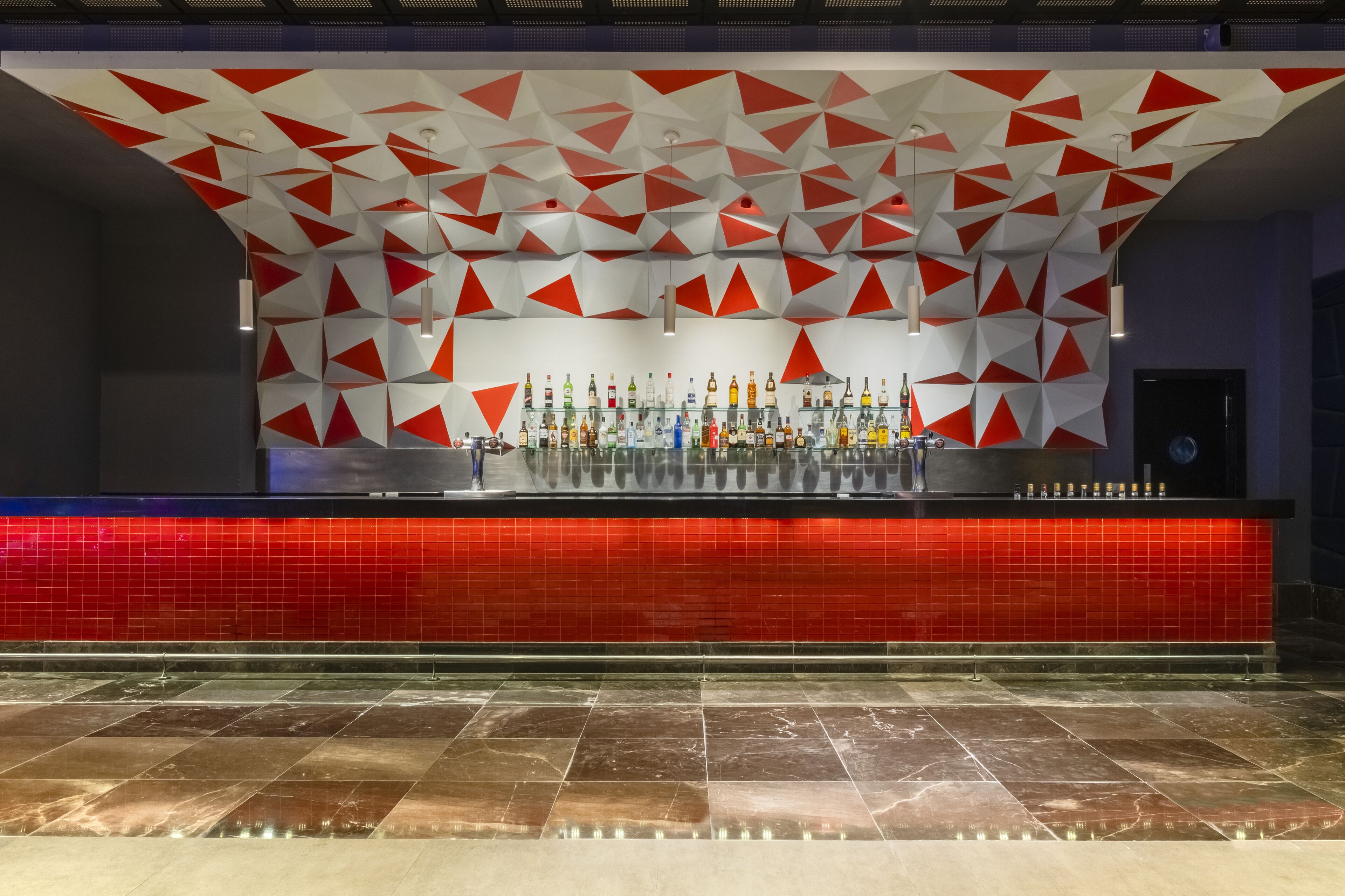 A bar area with a red tiled backsplash, a geometric ceiling design, and a variety of bottles on the counter.