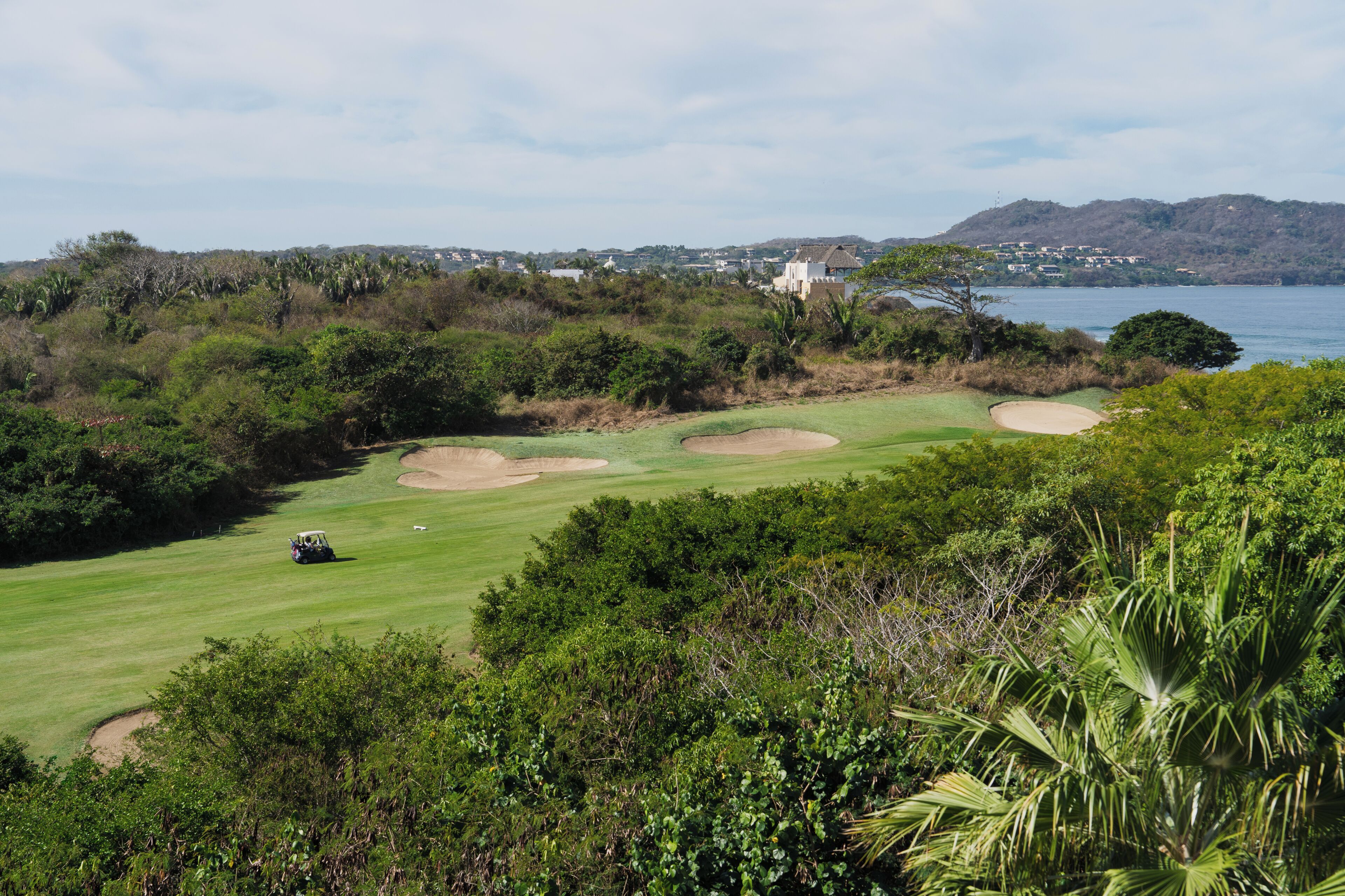 A golf course with bunkers, a water hazard, and a distant view of a coastal town.